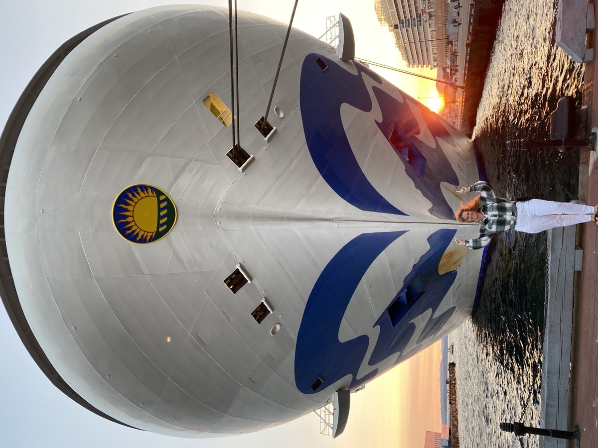 Liz standing beneath the towering bow of a cruise ship at sunset